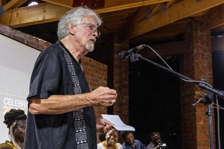 Un homme aux cheveux et à la barbe grisonnants parle devant un microphone, une rangée de personnes assises derrière lui.