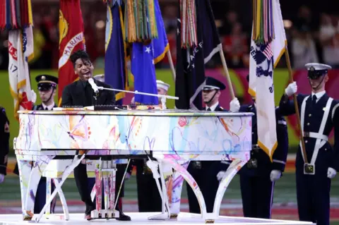 Getty Images Jon Batiste est assis dans un piano à queue Technicolor, flanqué de membres de l'US Air Force, qui tiennent des drapeaux