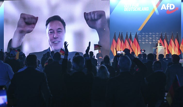 Homme avec des poings surélevés sur un écran lors d'un grand rassemblement avec beaucoup de drapeaux allemands.