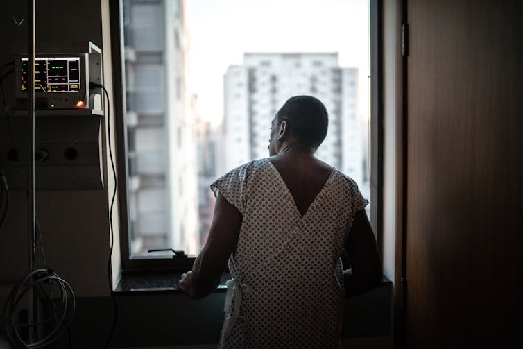 Patient en robe d'hôpital regardant par fenêtre, mains légèrement contreventement contre le seuil