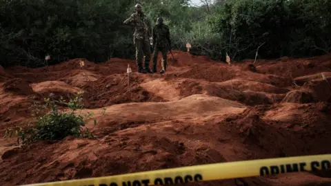 AFP via Getty Images Deux hommes en uniforme militaire se tiennent sur de la terre rouge où plusieurs grands trous ont été creusés. Derrière eux, on aperçoit des arbres. Au premier plan se trouve une bannière jaune sur laquelle on peut lire : La scène du crime ne traverse pas