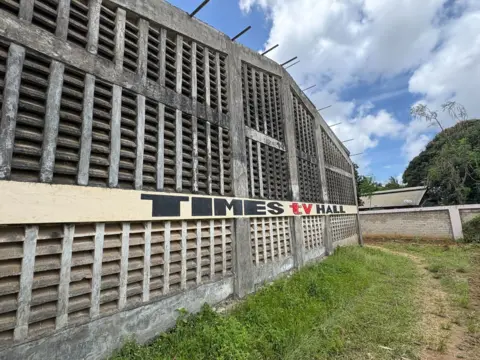 Un bâtiment en béton gris avec de nombreuses bouches d'aération dans le mur. Il y a un panneau indiquant Times TV Hall