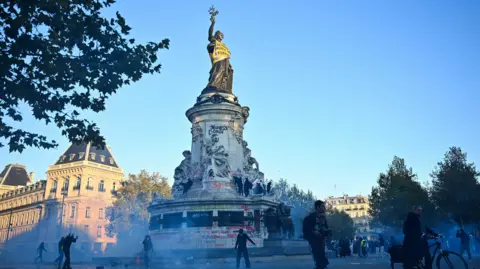AFP via Getty Images Des manifestants marchent à côté des gaz lacrymogènes et de la « Statue de la République » alors qu'ils se rassemblent alors que des affrontements ont éclaté sur la place de la République à la suite d'une manifestation dans le centre de Paris, le 18 octobre 2026.